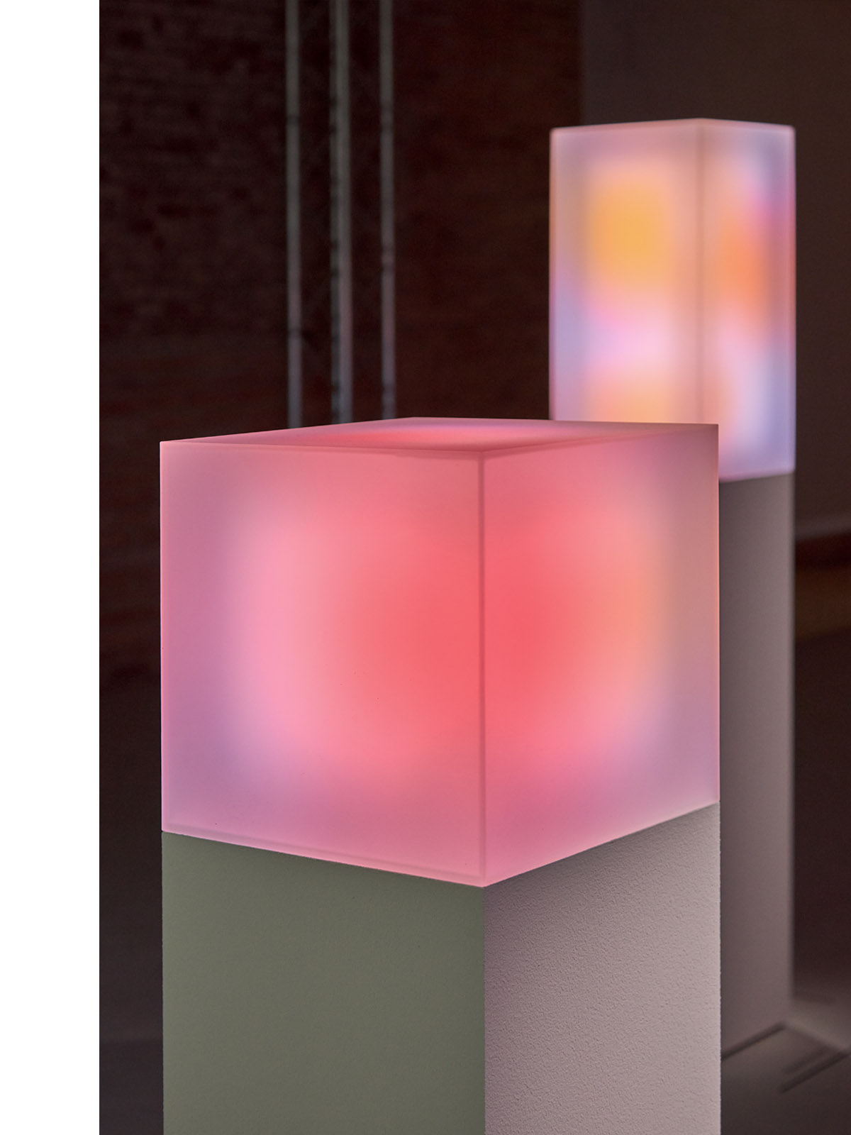 빛과 색에 중점을 둔 미술 설치의 일부로, 배경에 은은하게 빛나는 다채로운 큐브와 함께 받침대 위에 빛나는 분홍색 큐브의 클로즈업.