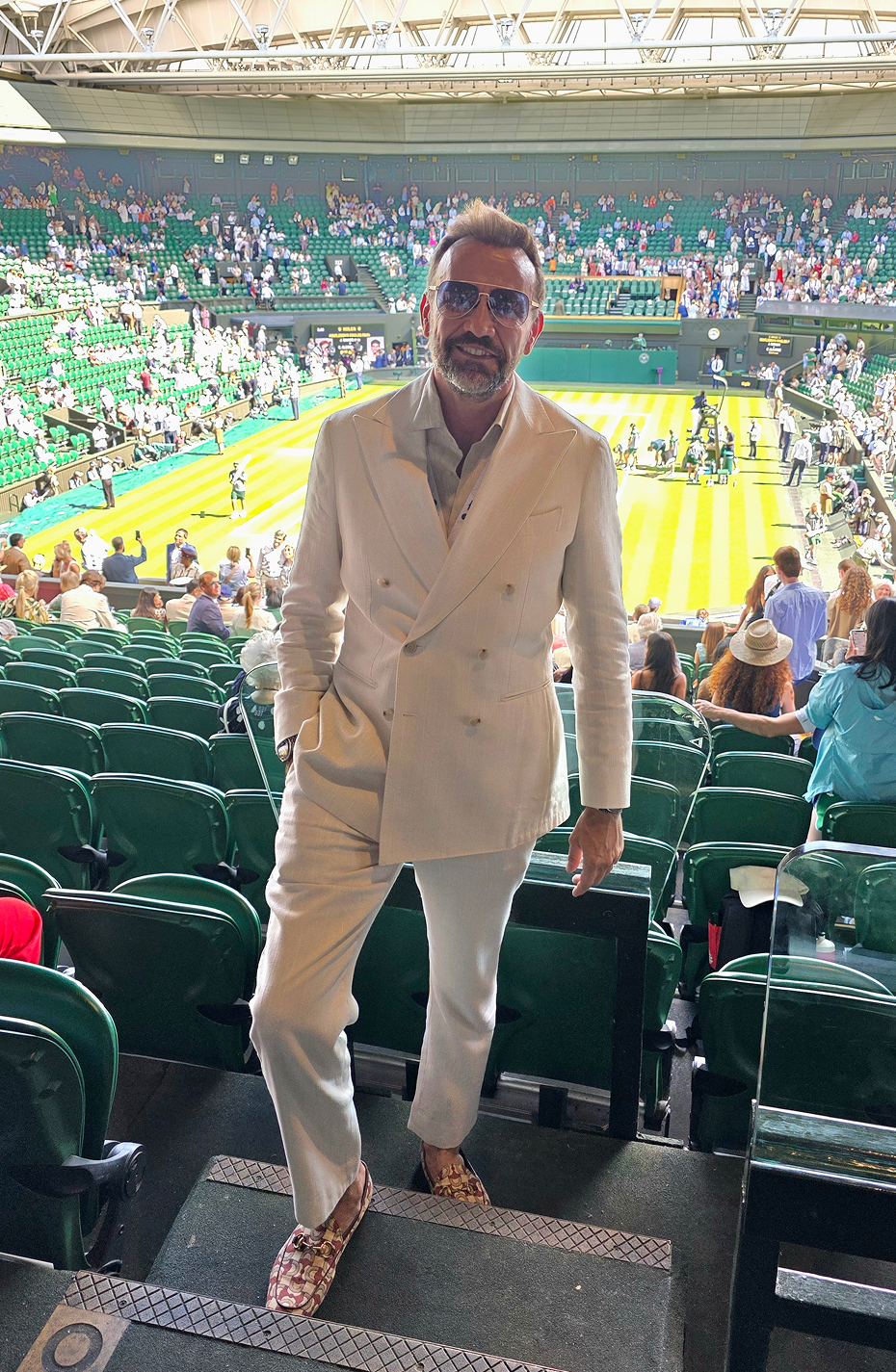 Against the backdrop of a tennis court, Mauro Porcini poses wearing a white set-up outfit and sunglasses, with one hand in his pants pocket.