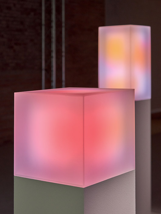 Close-up of a glowing pink cube on a pedestal with a softly lit, colorful cube in the background, part of an art installation with a focus on light and color