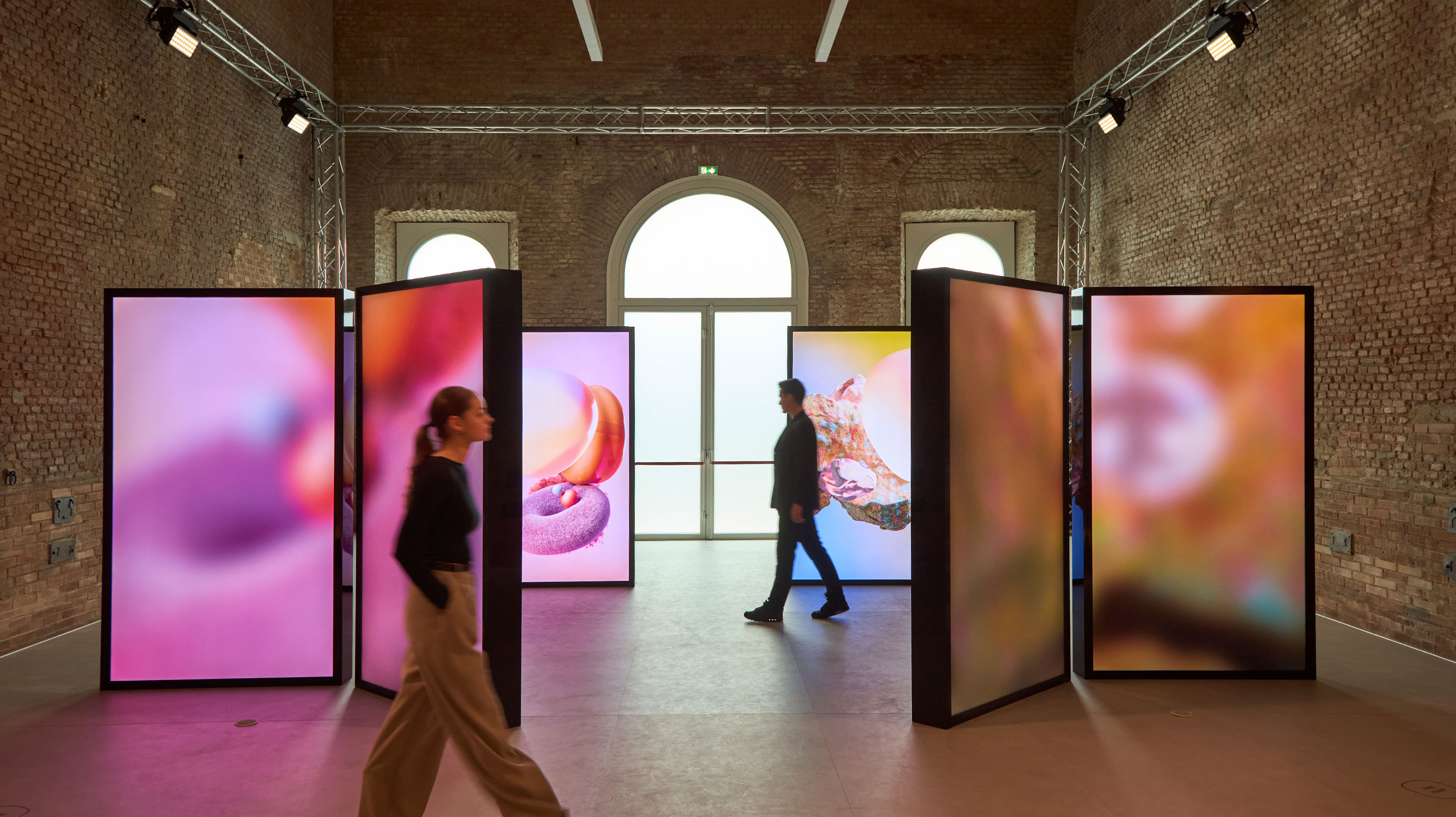 Visitors walking through an art exhibit featuring large, freestanding panels displaying vibrant abstract imagery, with arched windows in a room with exposed brick walls