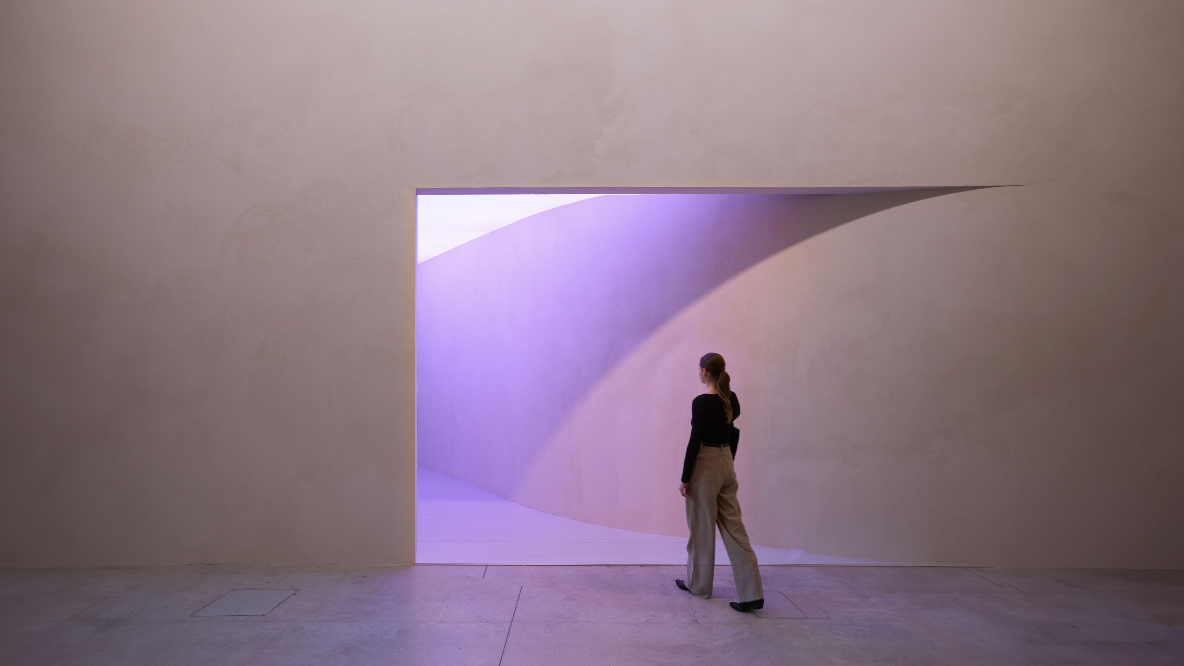 There is a door on a huge yellow wall, and purple light is leaking out. A young woman is walking toward the door.