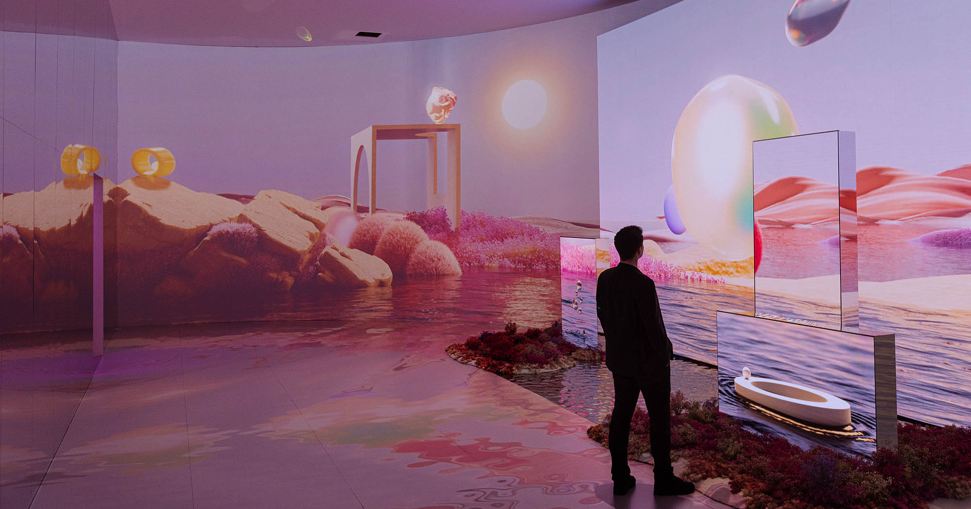 A person stands observing a large-scale, immersive art installation with surreal landscapes projected on walls and panels, creating an otherworldly atmosphere