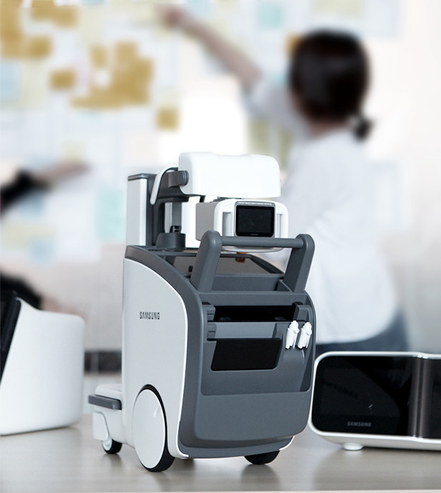 Medical devices and products are displayed on a table while a designer puts notes on a glass wall in the background.