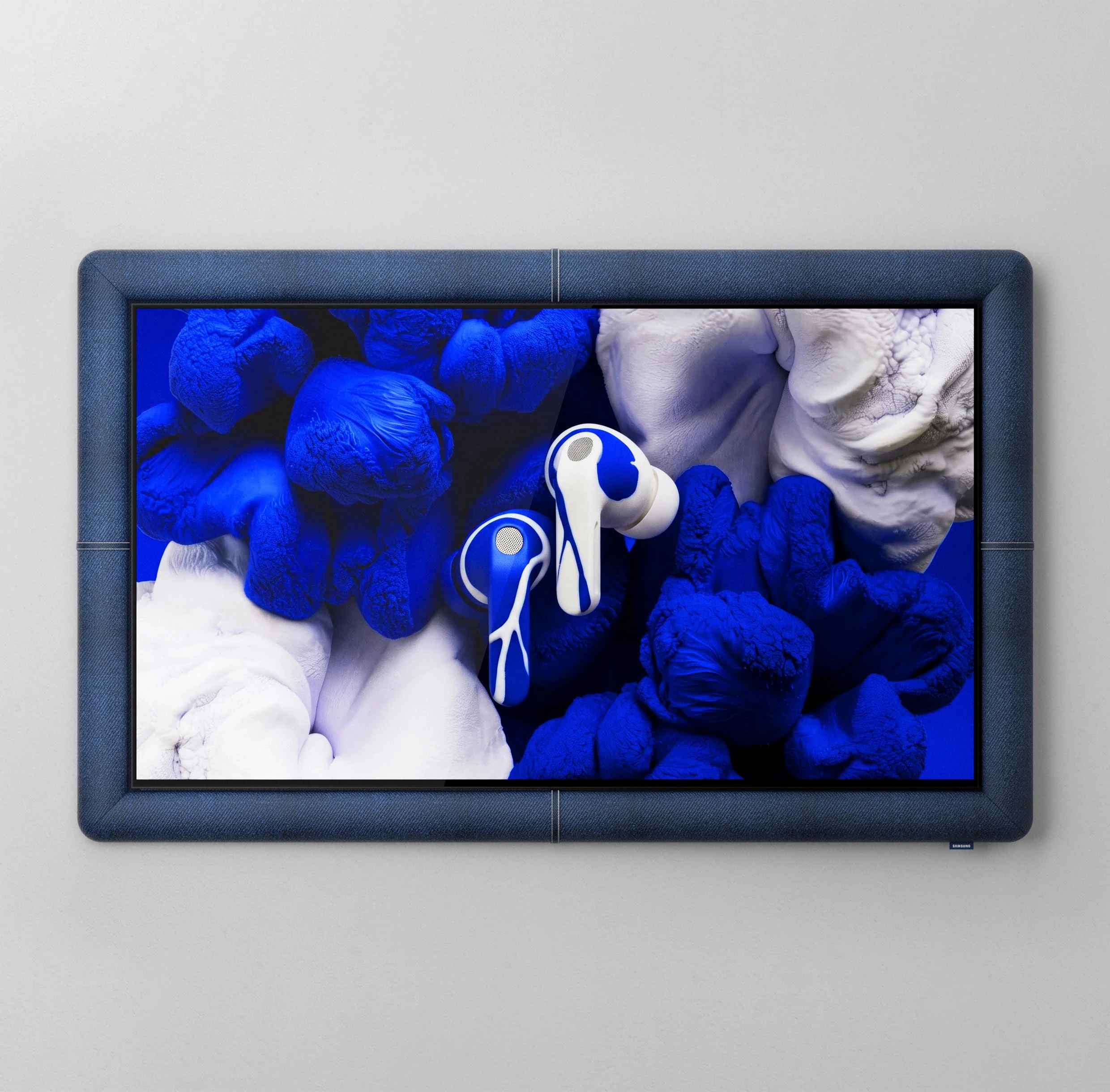 Two white Galaxy Buds4 Pro wireless earbuds float inside a horizontal frame wrapped in dark blue fabric. They are surrounded by blue and white folded fabrics and soft, rounded forms.
